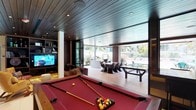 This single-floor space is designed as a high-end entertainment and social hub, combining multiple distinct zones within a cohesive, open layout. Rich dark wood ceiling planks run continuously throughout, complemented by recessed lighting that creates a warm, refined atmosphere across each area.

The entertainment zone anchors the space with a full-size red billiards table positioned on light-toned tile flooring. Adjacent to it, a media lounge features built-in dark wood shelving surrounding a flat-screen television, with deep brown leather sectional seating accented by dark satin cushions. A vibrant multicolored area rug and a sculptural gold accent chair add visual contrast and personality to the seating area.

A dramatic bar area showcases a backlit agate stone feature wall in amber and grey tones, serving as a striking focal point behind a dark wood bar counter with gold-accented stools. A glass partition separates this zone from a formal dining area, which is anchored by a round table beneath an ornate chandelier.

Floor-to-ceiling glass walls and sliding doors open the space toward an outdoor terrace and pool area, where a contemporary dining set and lush tropical landscaping are visible, flooding the interior with natural light and seamlessly blending indoor and outdoor living.
