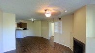 This single-floor residence offers a practical and comfortable layout designed for everyday living. The main living area is a spacious, open room finished with rich, dark hardwood-style flooring that extends throughout, creating a warm and cohesive aesthetic. A textured popcorn ceiling adds a classic touch, while a central ceiling light fixture provides ambient illumination. A notable feature of the living space is the built-in wall shelving unit, offering both display and storage opportunities, as well as a fireplace that serves as a natural focal point. The room is well-ventilated with visible HVAC vents integrated discreetly into the walls and ceiling. Adjacent to the living area, the kitchen is accessible via an open pass-through counter with bar-top seating, making it ideal for casual dining or entertaining. The kitchen features dark wood cabinetry that complements the flooring. A hallway leads further into the home, suggesting additional rooms beyond the main living space. Neutral, light-toned walls throughout provide a clean backdrop that makes the space feel bright and move-in ready. The overall ambiance is functional and inviting, with classic residential details throughout.