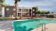 Located at 3055 N Flowing Wells Rd in Tucson, Arizona, this single-level outdoor amenity space offers a well-appointed recreational area designed for relaxation and leisure. The centerpiece of the space is a generously sized swimming pool with clear blue-green water, accessible via metal handrail entries and surrounded by a wide concrete pool deck that provides ample room for lounging. Adjacent to the pool sits a raised circular spa, finished with textured stonework, offering a dedicated space for hydrotherapy. The surrounding landscape features a mix of mature palm trees, shade trees, and manicured shrubs, adding greenery and a desert-friendly aesthetic to the environment. A distinctive architectural element of the space is the arched colonnade facade of the adjacent building, featuring decorative wrought iron railings and stucco detailing that lend a Southwestern character to the area. The multi-story residential building visible from the pool deck features arched balconies, exterior stairwells, and landscaped planters that frame the courtyard setting. The entire pool area is enclosed by a metal security fence, ensuring safety and privacy. Open skies above allow for abundant natural sunlight, making this outdoor space ideal for enjoying Tucson's warm climate year-round.