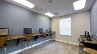 The computer lab is a focused, single-room workspace designed to provide a functional and comfortable environment for computer use. The room features a wall-mounted granite-style counter running along one side, hosting multiple desktop computer workstations positioned side by side, each paired with a modern wooden barstool with a chrome frame. This counter-height setup creates an efficient use of space while maintaining an organized, clean aesthetic.

The room is finished with a drop ceiling fitted with recessed fluorescent panel lighting, ensuring even, well-distributed illumination throughout the space. The walls are painted in a soft blue-gray tone, contributing to a calm and focused atmosphere. A window fitted with white horizontal blinds allows for natural light while maintaining privacy. The flooring consists of patterned carpet tiles in neutral tones, adding texture and comfort underfoot.

Overall, the computer lab is thoughtfully laid out to accommodate multiple users simultaneously, offering a practical and tidy setting suited for work, research, or general computer access.