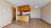 This single-floor unit offers a functional, straightforward layout designed for comfortable everyday living. The open-concept kitchen and living area serves as the heart of the space, where a peninsula with a granite-style countertop separates the kitchen from the carpeted living area. The kitchen is equipped with warm wood cabinetry providing ample upper storage, a white refrigerator, an electric range with overhead ventilation, and a sink. Stone-look tile flooring runs throughout the kitchen, transitioning to carpet in the living area. The spacious living room receives generous natural light through a large sliding glass door fitted with vertical blinds, creating a bright and welcoming atmosphere. A hallway leads to the bedroom, which features soft carpeting, a sliding wood-panel closet, and a window with vertical blinds that invites additional natural light. Baseboard heating is present throughout. The bathroom is fitted with white square tile surrounding the tub-shower combination, a matching tile floor, a vanity with wood cabinetry, and a large mirror. The unit maintains a clean, neutral palette with beige walls and warm wood tones throughout.