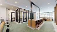 Located at 5905 Caravan Ct in Orlando, FL, this thoughtfully designed co-working and collaborative lounge spans a generous single floor, offering a variety of work and social environments within one cohesive space. The interior is anchored by a row of sleek, glass-enclosed private offices framed in bold black steel, providing visual transparency while maintaining acoustic separation for focused work. A long, warm-toned wood bar-height communal table runs alongside these offices, suspended pendant globe lights casting an inviting glow above it. The open lounge area features plush sectional seating upholstered in earthy tones and geometric patterns, complemented by small bistro tables and accent stools, creating an ideal setting for informal meetings or relaxed work sessions. Tucked into the layout are intimate acoustic booth nooks with upholstered banquette seating and pendant lighting, offering semi-private spaces for calls or focused tasks. The flooring transitions between lush green carpet and textured neutral area rugs, adding visual warmth and zonal definition throughout. Wall art with nature-inspired motifs adds a refined aesthetic touch. Large windows allow natural light to filter in, enhancing the bright, airy ambiance. A hallway connects to adjacent spaces, extending the overall flow of the layout.