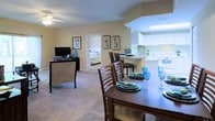 This single-story residence showcases an open-concept layout that seamlessly connects the main living areas, creating a spacious and cohesive environment throughout. The dining area serves as a central hub, accommodating a dark wood dining table with seating for multiple guests, elegantly set with decorative dishware and blue glassware that complement the home's cool-toned accents.

Adjacent to the dining space, the kitchen features white cabinetry extending to the upper walls, providing generous storage, along with white appliances and a peninsula breakfast bar that bridges the kitchen and dining areas. A skylight above the kitchen floods the space with natural light, enhancing the bright and airy atmosphere.

The living area flows naturally from the dining space and is finished with soft carpeted flooring. A ceiling fan with a light fixture provides both airflow and ambient lighting. The room comfortably accommodates entertainment furniture, including a media console with a flat-screen television.

Warm beige walls are accented by framed artwork, adding a polished, cohesive character throughout. A bedroom visible through an open doorway suggests a well-connected, practical floor plan ideal for comfortable everyday living.