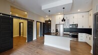 Located in Decatur, GA, this single-floor unit offers a thoughtfully designed open-concept layout that seamlessly connects the kitchen, dining, and living areas. The kitchen is a standout feature, showcasing white shaker-style cabinetry extending to the ceiling, a bold dark geometric tile backsplash, stainless steel appliances including a French door refrigerator and built-in microwave, and a central island with a dark quartz countertop and undermount sink. Stylish pendant lighting in a matte black finish adds a modern touch overhead. A built-in wine rack and ample cabinetry provide generous storage throughout. Wide-plank hardwood flooring flows throughout the main living areas, complemented by neutral warm-toned walls and clean white trim. The living area is bathed in natural light from large French doors that open to a private balcony, offering outdoor access and an airy feel. Dramatic dark five-panel barn-style and swing doors create striking contrast against the light interior palette. The bathroom features large-format tile flooring, white subway tile surrounding a soaking tub, a dark quartz vanity top with white cabinetry, and matte black hardware, maintaining the cohesive modern aesthetic found throughout the space.