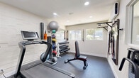 Located at 1039 N Alamo Ave in Tucson, Arizona, this dedicated exercise room offers a fully equipped fitness facility within a compact, efficiently designed space. The room features durable rubber flooring suitable for high-impact workouts, bright white walls — including a distinctive textured stone-accent wall — and recessed lighting throughout that creates a clean, energizing atmosphere.

The space is outfitted with a range of cardio and strength training equipment, including a treadmill with an integrated display, a stationary air bike, and a weight bench. A full rack of hex dumbbells in varying weights is mounted along one wall, alongside a tiered medicine ball rack and a stability ball, providing comprehensive free-weight training options. Resistance bands and suspension training handles are mounted to the wall, offering additional versatility for functional fitness.

Large windows along one wall allow natural light to filter in and provide views of the surrounding outdoor greenery, contributing to an open and inviting workout environment. A wall-mounted fan supports ventilation and comfort during exercise. A paper towel dispenser and cleaning supplies are conveniently accessible, maintaining hygiene standards. The room's layout is thoughtfully arranged to maximize usability, and a glazed door connects the fitness space to an adjacent lounge area, making it a well-integrated amenity within the broader facility.