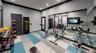 The Spin Room is a purpose-built fitness space designed to support a dynamic and comprehensive workout experience. The room features a row of stationary spin bikes positioned along a full-length mirrored wall, which creates a sense of depth and allows cyclists to monitor their form throughout a session. Integrated flat-screen televisions mounted on the walls display on-demand workout content, enhancing the interactive fitness experience. The flooring features a bold blue, white, and gray checkered tile pattern that adds visual energy to the space while providing a durable, easy-to-maintain surface. Complementing the cycling equipment, the room also includes a dumbbell rack stocked with a full range of free weights, a medicine ball rack with various weighted balls, a fitness bench, and an Elevate punching bag, making it suitable for cross-training and strength conditioning as well as cardio. The walls are finished in a cool neutral gray with dark crown molding trim, lending the space a clean, modern aesthetic. Recessed lighting provides bright, even illumination throughout. A posted set of Spin Rules on the wall ensures a structured and respectful environment for all users. The room is accessed through dark-framed doors, reinforcing the sleek, contemporary design of the overall space.