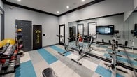 The Spin Room is a purpose-built fitness space designed to support a dynamic and comprehensive workout experience. The room features a row of stationary spin bikes positioned along a full-length mirrored wall, which creates a sense of depth and allows cyclists to monitor their form throughout a session. Integrated flat-screen televisions mounted on the walls display on-demand workout content, enhancing the interactive fitness experience. The flooring features a bold blue, white, and gray checkered tile pattern that adds visual energy to the space while providing a durable, easy-to-maintain surface. Complementing the cycling equipment, the room also includes a dumbbell rack stocked with a full range of free weights, a medicine ball rack with various weighted balls, a fitness bench, and an Elevate punching bag, making it suitable for cross-training and strength conditioning as well as cardio. The walls are finished in a cool neutral gray with dark crown molding trim, lending the space a clean, modern aesthetic. Recessed lighting provides bright, even illumination throughout. A posted set of Spin Rules on the wall ensures a structured and respectful environment for all users. The room is accessed through dark-framed doors, reinforcing the sleek, contemporary design of the overall space.