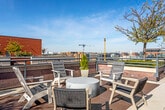 This rooftop terrace is a well-appointed outdoor amenity space designed to offer both relaxation and entertainment within an urban setting. The terrace is defined by warm red brick architectural walls that frame the space and provide a sense of enclosure while preserving sweeping views of the surrounding cityscape and open sky.

The terrace is organized into several distinct zones. One area features a casual seating arrangement of mixed outdoor chairs surrounding a round side table, positioned to take full advantage of the panoramic views. A shade umbrella nearby adds practical comfort during sunny days.

A covered lounge area anchored by a wood-slat pergola provides a shaded retreat. Beneath it, a contemporary sofa set and low coffee tables face a wall-mounted outdoor television, making it well-suited for social gatherings. The pergola is backed by a striking red wood-paneled wall that adds warmth and architectural character.

A more open section of the terrace offers large-format paver tiles, planters with greenery, and space for grilling, with propane tanks and a grill visible nearby. Raised garden beds and ornamental trees are distributed throughout, softening the hardscape with natural elements.

Access to the terrace is provided through glass-and-metal doors framed by brick, connecting the outdoor space to the building interior. The overall atmosphere is airy, modern, and well-suited for urban outdoor living.