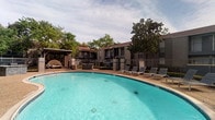 This outdoor pool area offers a well-maintained recreational space designed for relaxation and leisure. The centerpiece is a large, kidney-shaped swimming pool with clear turquoise water, surrounded by an aggregate concrete deck that provides ample space for movement and lounging. A row of sling-style lounge chairs lines one side of the pool, offering spots to sunbathe or unwind. A covered pergola structure at one end of the deck provides a shaded retreat, complementing the open-air environment.

The pool area is fully enclosed by a decorative wrought iron fence with brick pillar accents, ensuring both security and a defined boundary. Access is controlled through a gated entry, which features posted pool rules and a "No Lifeguard on Duty" sign for resident awareness. Safety equipment, including a life ring and an emergency call station, is mounted near the entrance gate.

Mature trees surrounding the perimeter provide natural shade and a sense of privacy. A shade sail is also installed in one section, offering additional sun protection. The overall setting is open-air and inviting, framed by the surrounding building exterior and lush landscaping with red mulch beds and greenery.