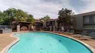 This outdoor pool area offers a well-maintained recreational space designed for relaxation and leisure. The centerpiece is a large, kidney-shaped swimming pool with clear turquoise water, surrounded by an aggregate concrete deck that provides ample space for movement and lounging. A row of sling-style lounge chairs lines one side of the pool, offering spots to sunbathe or unwind. A covered pergola structure at one end of the deck provides a shaded retreat, complementing the open-air environment.

The pool area is fully enclosed by a decorative wrought iron fence with brick pillar accents, ensuring both security and a defined boundary. Access is controlled through a gated entry, which features posted pool rules and a "No Lifeguard on Duty" sign for resident awareness. Safety equipment, including a life ring and an emergency call station, is mounted near the entrance gate.

Mature trees surrounding the perimeter provide natural shade and a sense of privacy. A shade sail is also installed in one section, offering additional sun protection. The overall setting is open-air and inviting, framed by the surrounding building exterior and lush landscaping with red mulch beds and greenery.