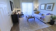 This single-floor residence offers a thoughtfully designed and elegantly appointed living environment. The living room serves as the centerpiece of the space, showcasing warm wood-look flooring that grounds the room with a sense of natural texture and continuity. A large sliding glass door fitted with vertical blinds and sheer curtains floods the room with abundant natural light, creating an open and airy atmosphere while providing access to an outdoor area beyond.

The living room is furnished with a cohesive and curated aesthetic. A plush white sofa accented with gold and patterned throw pillows anchors one side of the room, while a classic dark wood armchair adds contrast and character. A tufted ottoman with ornate turned legs sits at the center atop a large, intricately patterned area rug that defines the seating arrangement and adds visual depth to the space. A dark wood console along the wall provides both storage and display opportunities, complemented by tasteful wall art and soft ambient lighting from table lamps. The overall ambiance is warm, inviting, and refined, blending traditional furnishing details with a light, contemporary color palette that makes the space feel both functional and stylish.