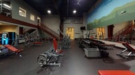 Located at 10 Newbridge Pkwy in Woodfin, NC, this fitness facility offers a spacious, single-floor layout designed to accommodate a comprehensive range of workout needs. The main training floor is generously proportioned, with soaring ceilings and a dark, industrial-style overhead finish that creates a focused, high-energy atmosphere. Ceiling fans and overhead lighting ensure the space remains well-lit and comfortable throughout.

A prominent feature of the facility is a large, vibrant mural depicting a rolling mountain landscape, which spans a significant portion of one wall and adds a distinctive local character to the environment. The rubber-floored training area is densely equipped with a full lineup of plate-loaded and selectorized strength machines, free weight stations, and an extensive dumbbell rack, all finished in a bold red-and-silver color scheme.

A red steel staircase leads to a mezzanine level with metal railing, visible from the main floor, adding vertical dimension to the space. Additional amenities include a front desk or check-in area, bulletin boards, built-in shelving, and a dedicated entry vestibule. Mounted televisions and an efficient layout make this facility a functional and well-organized training environment suited for serious fitness enthusiasts.
