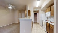 This single-floor residence is designed with an open-concept layout that seamlessly connects the kitchen and living areas. The kitchen is equipped with white appliances, including a refrigerator and range, complemented by warm oak-toned cabinetry that provides ample storage. A peninsula counter serves as a functional divide between the kitchen and the adjoining living space, offering additional workspace and a casual dining or gathering area.

The kitchen flooring features light-toned vinyl tile, which transitions into wall-to-wall carpeting in the living area, adding warmth underfoot. Neutral beige walls throughout the space create a cohesive, understated aesthetic. Overhead, a fluorescent light fixture in the kitchen ensures bright, practical illumination, while a ceiling fan in the living area provides both lighting and air circulation.

A hallway visible from the kitchen leads to additional rooms, with a glimpse of a laundry area featuring a stacked or side-by-side washer and dryer unit. The front entry door is visible from the living area, reinforcing the straightforward and accessible flow of the floor plan. Overall, the space presents a clean, functional environment suited to everyday living.