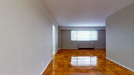 Located at 515 Mt Prospect Ave in Newark, NJ, this single-floor unit offers a functional and well-maintained layout with distinct living, kitchen, and bathroom areas. The main living space is a standout feature, showcasing richly toned, glossy parquet wood flooring arranged in a classic block pattern that adds warmth and character throughout. Neutral gray walls provide a clean, versatile backdrop, while a wide window fitted with horizontal blinds allows natural light to fill the room. A baseboard radiator unit runs along the exterior wall, providing efficient heating. The layout revealed in the dollhouse view shows a spacious open living area that flows toward a separate kitchen and bathroom corridor. The kitchen is well-appointed with white cabinetry offering ample upper and lower storage, white appliances including a gas range, built-in microwave, and refrigerator, and a striking multicolored mosaic tile backsplash that adds a decorative focal point. White floor tiles keep the kitchen bright and easy to maintain. The bathroom continues the clean aesthetic with white wall tiles, a tub-shower combination, a dark-finished vanity with under-sink storage, a mirrored medicine cabinet, and white floor tiles throughout. The overall ambiance is tidy, practical, and move-in ready.