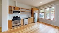 This single-story space is defined by warm hardwood flooring that runs consistently throughout, unifying the layout and lending a cohesive, natural aesthetic to every room. The open floor plan, as revealed in the overhead view, connects the main living areas in a straightforward, functional arrangement.

The kitchen is a standout feature, equipped with a full suite of stainless steel appliances including a refrigerator, electric range, microwave, and dishwasher. Natural wood cabinetry — both upper open shelving and lower enclosed units — lines the walls, complemented by a classic white subway tile backsplash and light countertops. A window beside the refrigerator brings in natural light, enhancing the workspace.

The living and bedroom areas are bright and airy, each fitted with ceiling fans and windows that frame views of surrounding greenery and the neighborhood. The entry features a warm wood door with glass pane inserts, welcoming natural light into the main living space.

The bathroom maintains a clean, classic aesthetic with black-and-white hexagonal tile flooring, a pedestal sink, a mirrored medicine cabinet, and white walls. A hallway connects the rooms efficiently, maintaining an uncluttered flow throughout the space.