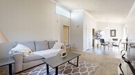 This efficient yet thoughtfully designed 1-bedroom, 2-bathroom single-floor residence offers a comfortable and functional layout. The open living and dining area is immediately welcoming, highlighted by striking vaulted ceilings with crown molding and a clerestory window that draws in natural light. Soft carpet underfoot and neutral tones throughout create a cohesive, calming ambiance. The living room provides ample space for relaxing, flowing seamlessly into the dining area, which is bathed in natural light from large windows and a sliding glass door that frames outdoor views of surrounding greenery. The kitchen features warm wood cabinetry, light stone-patterned countertops, and hardwood-style flooring, offering practical workspace and storage. The spacious primary bedroom is a comfortable retreat, complete with plush carpet, crown molding, a ceiling fan, and windows overlooking the outdoors. Two bathrooms serve the unit, one of which features a jetted soaking tub and shower combination, while the other offers a vanity with a wide mirror and hardwood-style flooring. Clean white walls and consistent finishes tie each space together, making this a well-rounded and inviting single-floor residence.