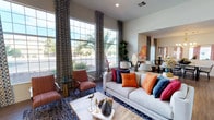 This single-story space showcases a thoughtfully designed open floor plan with a strong contemporary aesthetic. The living area is a standout, boasting soaring ceilings that amplify the sense of space, with floor-to-ceiling windows dressed in bold geometric-patterned drapery that frame views of lush outdoor palms and desert landscaping. A textured stacked-stone accent wall anchors a large flat-screen television, while wood-look flooring, a patterned area rug, and vibrant throw pillows in orange, red, and navy add warmth and personality. Oversized framed mirrors further enhance the room's openness.

The dining area flows naturally from the living space and features rich wood-look flooring, coordinating patterned curtains, and a striking mid-century modern sputnik chandelier in gold. A pass-through window connects this area to an adjacent space, creating seamless sightlines throughout. Bold wall art adds a colorful focal point.

The utility or laundry area is equally well-appointed, featuring dark wood cabinetry with brushed gold hardware, a deep stainless steel farmhouse sink with a matching gold faucet, crisp white countertops, and a dramatic navy elongated hexagon tile backsplash — a refined, detail-oriented finish that elevates even the most functional spaces.