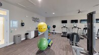 Located at 119 Dapple Ct in Wilmington, NC, this well-appointed fitness center is designed to support a comprehensive workout experience within a single, open-plan floor. The space features neutral gray walls, striped carpet flooring, and recessed lighting throughout, creating a clean and energizing atmosphere. Ceiling fans are distributed across the main workout area to maintain comfort during exercise sessions.

The cardio zone is equipped with multiple treadmills and elliptical machines from reputable brands including TRUE Fitness, each paired with individual display consoles. A bank of wall-mounted flat-screen TVs faces the cardio equipment, allowing members to stay entertained during workouts. The strength training area includes a multi-station cable machine, a dedicated free weights rack with a full dumbbell selection, adjustable benches, and a weight rack for a complete resistance training experience.

A functional fitness area houses an assortment of stability balls, medicine balls, and yoga mats organized on a vertical storage rack. A vibrant teal accent wall with playful branding adds a pop of color to this zone. A small refreshment counter with a compact refrigerator is conveniently located nearby. Natural light filters in through multiple windows along one wall, and a glass-panel entry door provides a welcoming entrance to the facility.