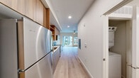 This contemporary studio in Burien, Washington offers a thoughtfully designed single-floor layout that efficiently combines living, kitchen, and bathroom spaces. Light hardwood flooring runs throughout, creating a warm and cohesive aesthetic from entry to living area.

The galley-style kitchen is a standout feature, equipped with a full suite of stainless steel appliances including a refrigerator, range, microwave, and dishwasher. Sleek gray lower cabinets are paired with rich wood-tone upper cabinetry and open shelving, complemented by white quartz countertops. Recessed lighting above keeps the space bright and functional.

The open living area benefits from large windows and a sliding glass door that leads to a private balcony, inviting abundant natural light and views of the surrounding community. A ceiling fan adds comfort to the space. The clean white walls and light flooring enhance the airy, modern ambiance throughout.

The full bathroom is finished with the same light hardwood-style flooring, a tub-shower combination, a modern floating vanity with a white countertop, and brushed nickel hardware accents. In-unit washer hookups are also present, adding practical convenience to this efficiently designed studio.