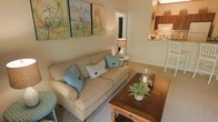 This single-floor residence offers a thoughtfully arranged layout with an open connection between the living area and kitchen. The living room centers around a cream-colored sofa with coral piping detail, accented by teal and white throw pillows that add a refreshing pop of color. A solid wood coffee table with turned legs serves as a focal point, styled with a small plant and decorative accessories. Above the sofa, a pair of botanical watercolor artworks in soft yellow, green, and orange hues complement the warm, neutral wall tones throughout. Soft carpeting underfoot adds comfort to the living space, while a side table with a table lamp provides warm ambient lighting. The open pass-through to the kitchen reveals dark wood cabinetry, a white range with overhead microwave, and a raised breakfast bar, offering both functionality and visual connection between spaces. A doorway visible in the background suggests additional rooms beyond, contributing to the unit's practical single-floor flow. The overall ambiance is warm, intimate, and well-curated, making it a welcoming and livable space.