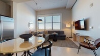 This thoughtfully designed single-floor residence offers a cohesive blend of contemporary style and everyday functionality. The open-concept living and dining area serves as the heart of the space, where a plush sectional sofa anchors the lounge zone beneath warm pendant lighting, complemented by a wall-mounted flat-screen TV and an arc floor lamp. Large windows dressed with horizontal blinds fill the area with natural light while providing adjustable privacy. A round dining table with modern black and blue chairs creates a distinct yet seamlessly connected dining area, accented by a circular wall mirror and a sculptural bowl centerpiece. The kitchen is efficiently laid out with two-tone cabinetry, stainless steel appliances including a range, dishwasher, and built-in microwave, all finished with under-cabinet lighting and clean countertops. The bedroom is a calm retreat featuring generous window coverage for natural light, a neatly made bed with crisp white linens, a teal upholstered bench, and a minimalist lounge chair. The bathroom is sleek and functional, offering a wide vanity with wood-tone cabinetry, a white countertop, and a glass-enclosed walk-in shower with mosaic tile flooring. Throughout the space, a neutral palette and consistent modern finishes create a refined and welcoming ambiance.