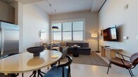 This thoughtfully designed single-floor residence offers a cohesive blend of contemporary style and everyday functionality. The open-concept living and dining area serves as the heart of the space, where a plush sectional sofa anchors the lounge zone beneath warm pendant lighting, complemented by a wall-mounted flat-screen TV and an arc floor lamp. Large windows dressed with horizontal blinds fill the area with natural light while providing adjustable privacy. A round dining table with modern black and blue chairs creates a distinct yet seamlessly connected dining area, accented by a circular wall mirror and a sculptural bowl centerpiece. The kitchen is efficiently laid out with two-tone cabinetry, stainless steel appliances including a range, dishwasher, and built-in microwave, all finished with under-cabinet lighting and clean countertops. The bedroom is a calm retreat featuring generous window coverage for natural light, a neatly made bed with crisp white linens, a teal upholstered bench, and a minimalist lounge chair. The bathroom is sleek and functional, offering a wide vanity with wood-tone cabinetry, a white countertop, and a glass-enclosed walk-in shower with mosaic tile flooring. Throughout the space, a neutral palette and consistent modern finishes create a refined and welcoming ambiance.