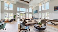 Located at 10491 Gandy Blvd N in Saint Petersburg, Florida, this single-floor community clubroom is a well-appointed, open-concept gathering space designed for both relaxation and recreation. The room immediately impresses with its dramatically soaring ceilings accented by exposed wood beams, while floor-to-ceiling windows and glass double doors flood the interior with abundant Florida sunshine and offer views of the outdoor surroundings.

The light hardwood flooring runs throughout, complementing the neutral gray-and-white color palette that gives the space a clean, contemporary feel. A generous lounge area anchors one side of the room, furnished with a plush sectional sofa, leather accent chairs, and a round coffee table — ideal for casual conversation or unwinding. Adjacent to this is a ping-pong table and a large flat-screen TV mounted above a built-in media console, catering to entertainment and social activities.

A well-equipped kitchenette features sleek gray cabinetry, a geometric mosaic tile backsplash, open wood shelving, a stainless steel refrigerator, microwave, and a black matte sink with white quartz countertops. A communal dining and work table with bar-height seating rounds out the space. A dartboard adds an extra recreational touch. The overall ambiance is bright, airy, and inviting — a versatile space suited for gatherings, work, or leisure.