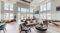 Located at 10491 Gandy Blvd N in Saint Petersburg, Florida, this single-floor community clubroom is a well-appointed, open-concept gathering space designed for both relaxation and recreation. The room immediately impresses with its dramatically soaring ceilings accented by exposed wood beams, while floor-to-ceiling windows and glass double doors flood the interior with abundant Florida sunshine and offer views of the outdoor surroundings.

The light hardwood flooring runs throughout, complementing the neutral gray-and-white color palette that gives the space a clean, contemporary feel. A generous lounge area anchors one side of the room, furnished with a plush sectional sofa, leather accent chairs, and a round coffee table — ideal for casual conversation or unwinding. Adjacent to this is a ping-pong table and a large flat-screen TV mounted above a built-in media console, catering to entertainment and social activities.

A well-equipped kitchenette features sleek gray cabinetry, a geometric mosaic tile backsplash, open wood shelving, a stainless steel refrigerator, microwave, and a black matte sink with white quartz countertops. A communal dining and work table with bar-height seating rounds out the space. A dartboard adds an extra recreational touch. The overall ambiance is bright, airy, and inviting — a versatile space suited for gatherings, work, or leisure.