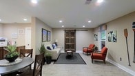 Located at 1053 Rifle Range Rd in Mt. Pleasant, South Carolina, this single-floor space offers a thoughtfully designed interior that balances rustic charm with modern comfort. The main living area serves as the centerpiece of the space, featuring light wood-toned flooring that enhances the bright, airy atmosphere created by recessed ceiling lights and natural light filtering through windows with colorful valances. A striking rustic sliding barn door adds a distinctive architectural focal point, complementing the neutral warm-toned walls throughout. The living area is furnished with a comfortable light-toned sofa accented with blue throw pillows, paired with vibrant burnt-orange accent chairs and glass-topped side tables, creating an inviting and cohesive aesthetic. Framed artwork adorns the walls, adding visual interest and a personalized touch to the space. A dining area is situated adjacent to the living space, with dark wood chairs visible, reinforcing the space's blend of rustic and contemporary design elements. Lush potted greenery is thoughtfully placed throughout, bringing a natural, refreshing quality to the interior. Overall, the space presents a warm, welcoming ambiance ideal for both relaxing and gathering.