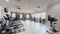 This single-level fitness center offers a generous, open layout designed to accommodate a wide range of workout styles. The space is finished with light wood-look flooring throughout, lending a clean and modern aesthetic that contrasts effectively with the distinctive textured stone accent walls, which serve as a defining architectural feature across multiple zones of the facility.

The cardio area is equipped with a row of treadmills positioned along a windowed wall, allowing users to enjoy outdoor views and natural light during workouts. Ceiling fans with brushed gold fixtures are installed throughout, providing both ventilation and a refined visual detail. Recessed lighting and crown molding add to the polished interior finish.

A strength training section houses a multi-station cable machine and several selectorized weight machines, while a dedicated free weights area includes a dumbbell rack, adjustable benches, and medicine balls. TRX suspension trainers are anchored in a functional training zone, complemented by a stacked stone half-wall partition. Stationary bikes occupy their own open area, offering additional cardio options.

Large windows throughout invite abundant natural light, and a wall of mirrors in the free weights area enhances the sense of space. A seating bench near the entrance and clearly marked exit doors complete the practical, well-considered layout.