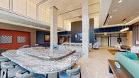 This upscale amenity lounge is thoughtfully designed to offer a dynamic and comfortable communal experience. The space is anchored by a striking curved granite bar countertop with brushed metal edging, complemented by a sleek cabinetry wall accented with vertical navy blue slat paneling and an integrated television — ideal for social gatherings. Soaring double-height ceilings lend a grand sense of openness throughout, while large floor-to-ceiling windows flood the interior with abundant natural light and frame views of the surrounding landscaped environment. The lounge is divided into several distinct seating areas, each furnished with curated, colorful pieces — including teal velvet sofas, jewel-toned armchairs in purple and blue, and patterned area rugs — creating an inviting and vibrant atmosphere. A dedicated recreational zone features dual shuffleboard tables set against a blue upholstered accent wall adorned with original artwork, while a bold terracotta-red accent wall displays oversized decorative chess boards. Additional flexible seating and bistro-style high-top tables positioned near the windows offer views and casual gathering spots. The polished stone flooring and recessed lighting throughout contribute to the space's refined, contemporary aesthetic.
