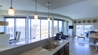 This single-floor urban residence offers a seamlessly open layout that maximizes both space and natural light. The kitchen serves as a central anchor, featuring a granite countertop island with an undermount sink and rich dark cabinetry. Pendant lights hang above the island, providing warm, focused illumination that complements the natural light pouring in from the surrounding floor-to-ceiling windows. These expansive windows curve gracefully along the perimeter, offering panoramic views of the surrounding cityscape — a standout architectural feature that creates a dramatic sense of openness and connection to the urban environment. The living area flows naturally from the kitchen, showcasing hardwood flooring and ample space for lounge seating. A wall-mounted flat-screen television is positioned to take advantage of the open plan, while comfortable armchairs are thoughtfully arranged to enjoy both the screen and the sweeping city views beyond the glass. The overall ambiance is modern and refined, blending functional design with upscale finishes. The abundance of natural light, city views, and cohesive open-plan design make this a distinctive and inviting urban living space.