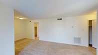 Located at 1518 Village Dr in Wilmington, North Carolina, this single-floor residence offers a clean and practical layout suited for comfortable everyday living. The main living area is finished with wall-to-wall carpet in a neutral beige tone, providing a warm underfoot feel throughout the space. The walls are painted in a crisp white, lending a bright and open atmosphere that makes the room feel airy and welcoming. Ceiling-mounted lighting provides consistent illumination, while HVAC vents positioned both on the ceiling and along the lower wall ensure efficient climate control year-round. A wall-mounted thermostat is conveniently accessible within the living area. A doorway visible from the main room leads to an adjacent space with warm-toned walls, suggesting a connected hallway or secondary room that adds to the overall flow of the floor plan. The textured ceiling adds a subtle architectural detail consistent throughout. Overall, the space presents a functional and well-maintained interior with a simple, neutral palette that offers a versatile canvas for a variety of furnishing and décor styles.