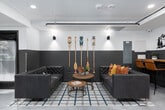 This single-level communal space showcases a contemporary design with a Pacific Northwest-inspired aesthetic throughout. The layout is thoughtfully organized into several distinct zones that flow seamlessly into one another, creating a versatile environment suited for both social gatherings and focused work.

The lounge area anchors the space with tufted leather sofas and upholstered sectional seating arranged atop a plaid area rug. Decorative oar paddles mounted vertically on a two-tone gray and white wainscoted wall serve as a striking focal point, complemented by mountain-themed metal wall art. Warm accent pillows in amber and orange tones add color throughout.

A large communal dining table crafted from natural wood accommodates generous seating beneath a trio of warm copper pendant lights, making it ideal for group meals or collaborative gatherings. Adjacent to the lounge, a smaller bar-height wood table with leather stools offers an informal workspace beneath exposed pipe runs along the ceiling.

The entry corridor features glass partitioning, safety signage, and polished concrete-style flooring that continues throughout the space, reinforcing the industrial yet welcoming character. Natural light filters in through street-facing windows fitted with roller shades. Recessed and wall-mounted lighting ensures the space is well-lit, while the overall design balances a rugged, nature-inspired warmth with clean, modern finishes.