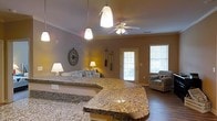 Located at 1001 Quarters Landing in Sneads Ferry, North Carolina, this single-story home offers a well-designed open-concept layout that seamlessly connects the kitchen and living areas. The kitchen is a standout feature, showcasing a generously sized curved granite countertop island that serves as both a functional workspace and a natural gathering point. Pendant lighting above the island adds a warm, inviting glow, complementing the ceiling fan that keeps the living space comfortable year-round. The living area is bright and airy, benefiting from natural light streaming through windows fitted with blinds, along with a glass-paned exterior door that enhances the sense of openness. Warm-toned walls and wood-style flooring run throughout, creating a cohesive and welcoming atmosphere. Coastal-inspired accents, including nautical wall décor and paddle ornaments, reflect the character of the surrounding Sneads Ferry community. The layout is thoughtfully arranged to maximize both flow and functionality, making this single-floor home an ideal retreat with a relaxed, comfortable ambiance suited to coastal living.