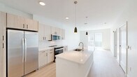 This well-appointed one-bedroom, one-bathroom unit at 100 Chestnut Street in Ashland, Massachusetts offers a thoughtfully designed open-concept layout with modern finishes throughout. The kitchen serves as the centerpiece of the living space, showcasing light wood cabinetry, white subway tile backsplash, stainless steel appliances—including a side-by-side refrigerator, electric range, and built-in microwave—and a large island with a quartz countertop and undermount sink, illuminated by stylish pendant lighting. Light-toned hardwood flooring flows throughout the main living area, enhancing the bright, airy ambiance. The bedroom features plush carpeting, large windows with blinds that invite natural light, and direct access to a dedicated closet. The bathroom is finished with a glass-enclosed walk-in shower clad in white subway tile and complemented by open wood shelving for added storage. A private balcony extends the living space outdoors, offering views of the well-maintained community courtyard and the surrounding multi-story residential buildings. The unit also includes a dedicated closet for additional storage. Clean white walls and contemporary hardware selections tie the space together with a cohesive, modern aesthetic.