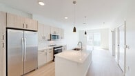 This well-appointed one-bedroom, one-bathroom unit at 100 Chestnut Street in Ashland, Massachusetts offers a thoughtfully designed open-concept layout with modern finishes throughout. The kitchen serves as the centerpiece of the living space, showcasing light wood cabinetry, white subway tile backsplash, stainless steel appliances—including a side-by-side refrigerator, electric range, and built-in microwave—and a large island with a quartz countertop and undermount sink, illuminated by stylish pendant lighting. Light-toned hardwood flooring flows throughout the main living area, enhancing the bright, airy ambiance. The bedroom features plush carpeting, large windows with blinds that invite natural light, and direct access to a dedicated closet. The bathroom is finished with a glass-enclosed walk-in shower clad in white subway tile and complemented by open wood shelving for added storage. A private balcony extends the living space outdoors, offering views of the well-maintained community courtyard and the surrounding multi-story residential buildings. The unit also includes a dedicated closet for additional storage. Clean white walls and contemporary hardware selections tie the space together with a cohesive, modern aesthetic.