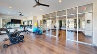 Located at 11335 N Michigan Rd in Carmel, Indiana, this fitness center offers a comprehensive and modern workout facility spread across a single floor. The space is divided into two distinct exercise rooms, each thoughtfully designed to accommodate a variety of fitness activities.

The main exercise area impresses with soaring double-height ceilings and an abundance of natural light streaming through floor-to-ceiling windows and upper clerestory windows, offering open views of the surrounding landscape. This expansive area houses a full lineup of cardio equipment, including treadmills, ellipticals, and stair climbers, complemented by a row of mounted televisions for entertainment during workouts. A water station is conveniently positioned nearby.

The second exercise room is separated by sleek glass partition walls, maintaining an open and airy feel while defining the space. This room features a variety of strength training machines, a dumbbell rack with an adjustable bench, full-length mirrors, and wall-mounted TVs. A dedicated studio area accommodates cycling bikes, medicine balls, resistance bands, and other functional fitness accessories, supported by a ceiling fan and a large wall-mounted display.

Throughout both rooms, warm hardwood-style flooring adds a refined aesthetic, while recessed lighting ensures a bright, motivating atmosphere. The overall design is clean, modern, and purpose-built for a high-quality fitness experience.