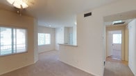 Located at 1390 Santa Alicia in Chula Vista, this single-floor unit features a clean, open layout designed to maximize usable living space. The interior is finished in crisp white walls paired with neutral-toned wall-to-wall carpeting, creating a light and airy atmosphere throughout. The living area is generously proportioned, with a window fitted with horizontal blinds that allows natural light to filter in, brightening the space. A central HVAC vent is integrated into the ceiling, ensuring comfortable climate control year-round. The kitchen is defined by a pass-through counter with a breakfast bar overhang, offering a functional transition between the cooking and living areas while maintaining an open, connected feel. Recessed lighting complements the natural light, keeping the interior well-illuminated. A paneled door visible beyond the kitchen counter leads to an additional area of the unit. Clean white baseboards frame the carpeted floors throughout, adding a polished finishing detail. The overall design emphasizes simplicity and functionality, making the space feel welcoming and move-in ready.