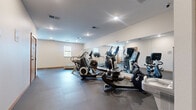 The fitness center at 3090 Hazelwood St in Maplewood, Minnesota, is a dedicated, single-room exercise facility designed to support a well-rounded workout experience. The space is laid out on a single floor and offers a comfortable, functional environment for residents or members seeking convenient access to fitness amenities.

The room features durable rubber tile flooring throughout, providing cushioning and stability for a variety of exercise activities. Recessed lighting illuminates the space evenly, while multiple windows along one wall admit natural light and offer views of the surrounding greenery, creating a bright and motivating atmosphere.

A large wall-mounted mirror spans a significant portion of one wall, enhancing the sense of openness and allowing users to monitor their form during workouts. The cardio equipment lineup includes treadmills, elliptical cross-trainers, and a stationary cycle, all from recognized fitness brands such as Star Trac and Life Cycle. A dedicated free weight area features a multi-tier dumbbell rack with a range of weights, accompanied by a weight bench and exercise mat for strength and flexibility training. A wall-mounted water station provides convenient hydration access.

The room's neutral-toned walls, clean lines, and organized layout contribute to a focused, no-frills fitness environment. A wood-trimmed entry door provides secure, straightforward access to the facility.
