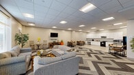Located at 3101 Euclid Ave in Cleveland, Ohio, this single-floor community space offers a generously sized, open-plan layout designed for comfort and functionality. The room is anchored by a bold chevron-patterned carpet that visually divides the lounge area from the kitchen, creating a dynamic and modern aesthetic throughout.

The lounge area is thoughtfully arranged with multiple seating groupings, including plush grey sofas, a checkered loveseat, and accent chairs positioned around coffee tables — ideal for relaxed gatherings or social interaction. A wall-mounted flat-screen television serves as a focal point for one of the seating arrangements, while tall decorative plants and warm accent pieces add a welcoming, lived-in feel. Natural light filters in through several windows fitted with blinds, brightening the neutral-toned walls and drop-panel ceiling overhead.

Transitioning from the lounge, the fully equipped kitchen features white shaker-style cabinetry, granite-look countertops, and a suite of black appliances including a range, refrigerator, and microwave. Light wood-look tile flooring grounds the kitchen area, offering a clean contrast to the carpeted lounge. A dining area with bar-height tables and chairs bridges the two zones seamlessly. This well-appointed space is well-suited for community use, resident amenities, or shared common area functions.