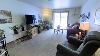 This single-floor space centers around a comfortable and well-appointed living room that combines functionality with a homey aesthetic. The room is carpeted throughout, providing a soft, warm underfoot feel that contributes to its inviting atmosphere. A large sliding glass door serves as a prominent architectural feature, allowing an abundance of natural light to fill the space while offering a direct view of the outdoors. The door also provides convenient access to the exterior.

The living room is thoughtfully arranged with multiple seating options, including a plush sofa adorned with decorative pillows and a matching armchair, both positioned to encourage relaxed conversation or media viewing. A round glass-topped coffee table anchors the seating area, while a wooden media console houses a flat-screen television along the opposite wall.

Lush indoor plants are distributed throughout the room, adding a natural, vibrant element to the décor and enhancing the overall sense of life and warmth within the space. A tall floor lamp provides warm ambient lighting, complementing the natural light from the sliding door. Decorative wall accents and personal touches give the space a lived-in, personalized character.