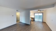 This single-floor space showcases an open-concept layout that seamlessly connects the living area and kitchen, creating a functional and cohesive living environment. The flooring throughout is finished in a warm wood-style material that adds continuity and a contemporary feel to the space.

The kitchen is partially separated by a half-wall partition, providing definition without sacrificing openness. It features white cabinetry, a stainless steel dishwasher, and a clean white tile backsplash. The design maximizes counter space and storage while maintaining a modern aesthetic.

The living area benefits from abundant natural light streaming through a large sliding glass door dressed with vertical blinds, which also provides direct access to an outdoor area. A ceiling fan with an integrated light fixture is positioned above, offering both air circulation and ambient lighting.

A hallway visible from the main living space leads toward additional rooms, suggesting practical separation of private areas. The walls are finished in a neutral light gray, complementing the flooring and overall palette. HVAC venting is integrated into the ceiling, and a thermostat is visible on the wall, indicating climate control throughout.