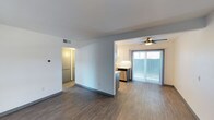 This single-floor space showcases an open-concept layout that seamlessly connects the living area and kitchen, creating a functional and cohesive living environment. The flooring throughout is finished in a warm wood-style material that adds continuity and a contemporary feel to the space.

The kitchen is partially separated by a half-wall partition, providing definition without sacrificing openness. It features white cabinetry, a stainless steel dishwasher, and a clean white tile backsplash. The design maximizes counter space and storage while maintaining a modern aesthetic.

The living area benefits from abundant natural light streaming through a large sliding glass door dressed with vertical blinds, which also provides direct access to an outdoor area. A ceiling fan with an integrated light fixture is positioned above, offering both air circulation and ambient lighting.

A hallway visible from the main living space leads toward additional rooms, suggesting practical separation of private areas. The walls are finished in a neutral light gray, complementing the flooring and overall palette. HVAC venting is integrated into the ceiling, and a thermostat is visible on the wall, indicating climate control throughout.