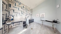 Located on Alexis Avenue in Daytona Beach, Florida, this purpose-built pet grooming space combines functional design with striking aesthetic details. The room's most prominent feature is a floor-to-ceiling accent wall clad in glossy geometric-patterned tile in white, gray, and teal — a bold visual centerpiece that sets an upscale tone throughout the space. Light gray large-format floor tiles provide a clean, durable surface underfoot, complete with a centrally located floor drain for easy cleanup. The grooming area is anchored by a stainless steel pet wash basin equipped with steps for easy pet access, alongside sleek dark navy cabinetry topped with white countertops that offer generous storage and workspace. A freestanding adjustable grooming table with harness attachments is positioned on the opposite side of the room, providing a dedicated area for styling and finishing. Natural light pours in through multiple windows fitted with horizontal blinds, and a glass-paneled entry door offers views of the surrounding exterior. The soaring ceiling height enhances the sense of openness within the compact footprint. Framed pet-themed artwork on the walls adds a charming, thematic touch, completing a well-designed grooming suite that is both practical and visually polished.