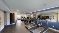 Located at 454 Heather Dr N in West Deptford, NJ, this fitness center offers a well-appointed, single-floor workout space designed to accommodate a comprehensive range of exercise needs. The room features rubber flooring throughout, providing durability and safety for all types of training activities.

The cardio section is prominently equipped with a row of TRUE-brand treadmills, stationary bikes, elliptical trainers, and rowing machines, all arranged for efficient use of the space. Large wall-mounted mirrors with integrated LED accent lighting line the main cardio wall, enhancing the sense of openness and allowing users to monitor their form during workouts.

The strength training area includes a multi-function cable machine, a dumbbell rack stocked with a full range of weights, and an adjustable workout bench. A wall-mounted flat-screen TV is positioned nearby for entertainment during training sessions.

Recessed ceiling lighting provides bright, even illumination throughout the space, complemented by natural light from windows along one wall. A bold motivational mural reading "Walk In Strong, Walk Out Stronger" serves as a focal point on one wall, reinforcing the energetic atmosphere. The color scheme of cool blue and neutral gray tones contributes to a modern, focused environment. The space is accessible via an interior door and features a glass-panel entry door that invites natural light from outside.