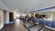 Located at 454 Heather Dr N in West Deptford, NJ, this fitness center offers a well-appointed, single-floor workout space designed to accommodate a comprehensive range of exercise needs. The room features rubber flooring throughout, providing durability and safety for all types of training activities.

The cardio section is prominently equipped with a row of TRUE-brand treadmills, stationary bikes, elliptical trainers, and rowing machines, all arranged for efficient use of the space. Large wall-mounted mirrors with integrated LED accent lighting line the main cardio wall, enhancing the sense of openness and allowing users to monitor their form during workouts.

The strength training area includes a multi-function cable machine, a dumbbell rack stocked with a full range of weights, and an adjustable workout bench. A wall-mounted flat-screen TV is positioned nearby for entertainment during training sessions.

Recessed ceiling lighting provides bright, even illumination throughout the space, complemented by natural light from windows along one wall. A bold motivational mural reading "Walk In Strong, Walk Out Stronger" serves as a focal point on one wall, reinforcing the energetic atmosphere. The color scheme of cool blue and neutral gray tones contributes to a modern, focused environment. The space is accessible via an interior door and features a glass-panel entry door that invites natural light from outside.
