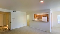 This single-floor space features an open-concept design that connects the living area to the kitchen through a convenient pass-through counter. The living area is finished with wall-to-wall carpet in a neutral beige tone, contributing to a clean and understated aesthetic. White walls throughout the space reflect light effectively, keeping the interior feeling bright and open. Recessed lighting in both the living area and kitchen provides consistent overhead illumination.

The kitchen is positioned at the rear of the main living space and is accessible via the pass-through counter, which also functions as a breakfast bar. The kitchen is equipped with warm-toned wooden cabinetry, a white refrigerator, and a sink centered beneath the upper cabinets. An under-cabinet range hood is also present, indicating a dedicated cooking area. The cabinetry provides ample storage, and the layout is compact yet functional.

A hallway visible from the living area leads to additional rooms, suggesting a practical floor plan that keeps the private spaces separated from the main living and kitchen areas. The overall ambiance of the space is practical and move-in ready.