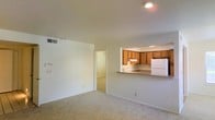 This single-floor space features an open-concept design that connects the living area to the kitchen through a convenient pass-through counter. The living area is finished with wall-to-wall carpet in a neutral beige tone, contributing to a clean and understated aesthetic. White walls throughout the space reflect light effectively, keeping the interior feeling bright and open. Recessed lighting in both the living area and kitchen provides consistent overhead illumination.

The kitchen is positioned at the rear of the main living space and is accessible via the pass-through counter, which also functions as a breakfast bar. The kitchen is equipped with warm-toned wooden cabinetry, a white refrigerator, and a sink centered beneath the upper cabinets. An under-cabinet range hood is also present, indicating a dedicated cooking area. The cabinetry provides ample storage, and the layout is compact yet functional.

A hallway visible from the living area leads to additional rooms, suggesting a practical floor plan that keeps the private spaces separated from the main living and kitchen areas. The overall ambiance of the space is practical and move-in ready.