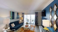 This single-floor, two-bedroom, two-bathroom residence offers a well-designed living space with a thoughtful blend of comfort and contemporary style. The living room serves as the centerpiece of the home, showcasing hardwood-style flooring, crown molding, and a bold, coordinated color scheme that ties the space together cohesively.

A striking navy blue tufted sofa anchors the seating area, complemented by a geometric marble-top coffee table and a vibrant orange-toned area rug that adds warmth and visual depth. A fireplace with a deep blue surround and mounted flat-screen television creates a compelling focal point on one wall, while an abstract canvas painting adds an artistic touch on the opposite side.

Natural light fills the room through a large sliding glass door dressed with patterned blue curtains, which opens directly onto a private balcony furnished with outdoor seating — ideal for relaxing or enjoying the surrounding views. The smooth white ceiling with crown molding detailing adds a clean, finished look throughout. Overall, the space delivers a vibrant yet inviting atmosphere suited for both everyday living and entertaining.